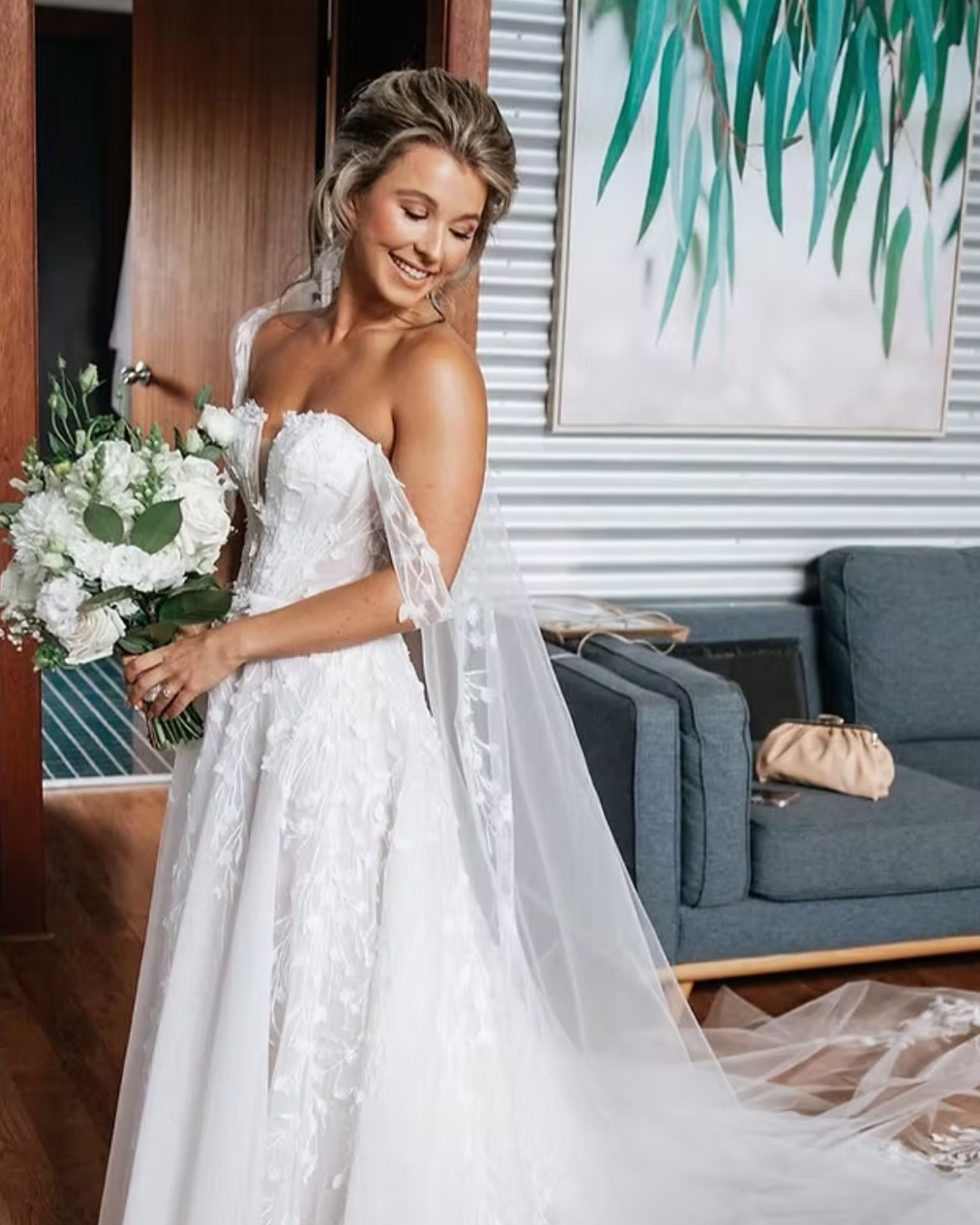 Woman in a white wedding dress holding a bouquet in a room with a couch and painting.