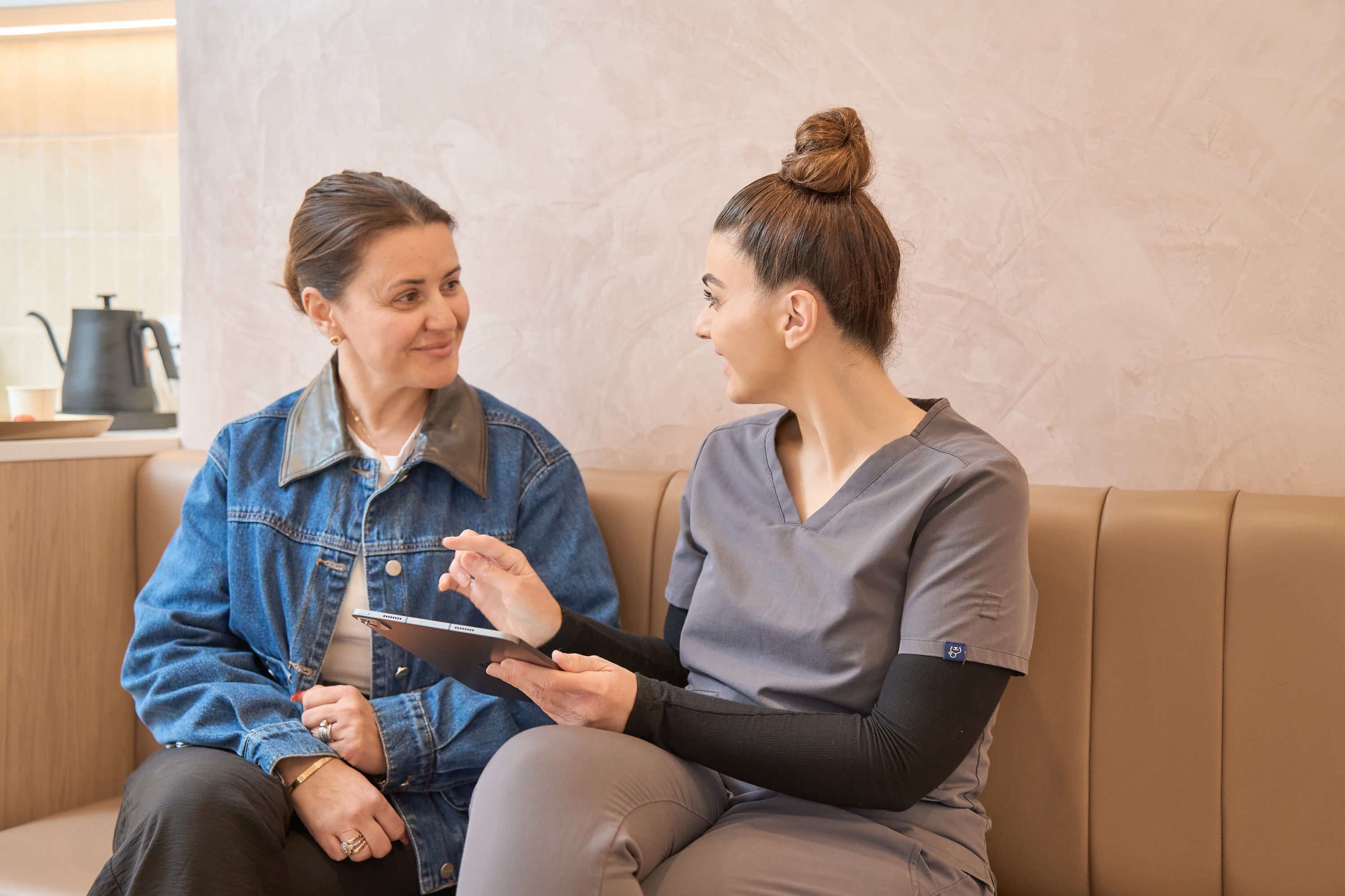 A woman in a blue denim jacket sits on a tan leather couch, smiling at a healthcare provider in gray scrubs who is holding a tablet and pen during a consultation.